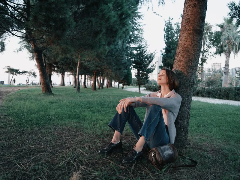 A lone person sits against a tree in a tranquil park, contemplating life. They are relaxed, leaning back, enjoying sunlight, shade, and a gentle breeze in a peaceful outdoor setting.