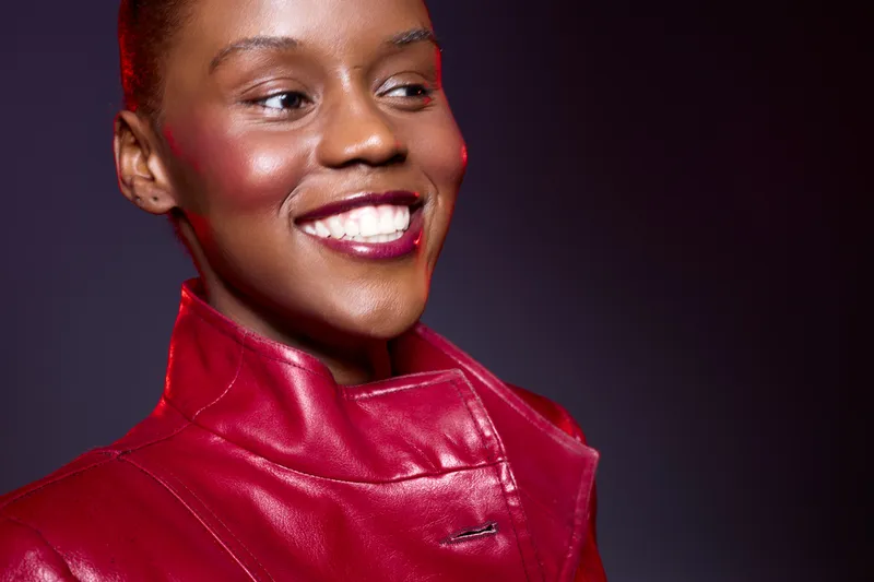 Close-up of a smiling black woman with dark skin wearing a red leather jacket against a dark purple background. Concept of fashion, beauty, confidence, and stylish clothing.