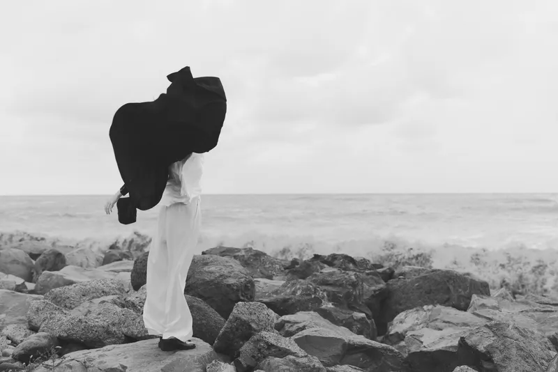 Monochrome silhouette walks along rocky shoreline, carrying a large coat over shoulder, sea mist in background, windy mood, dramatic coastal fashion scene.