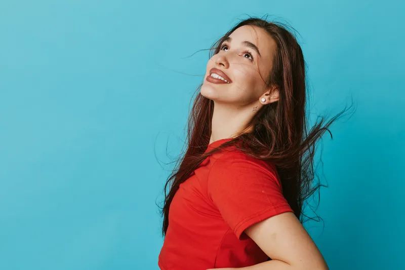 Young woman in red shirt smiling and looking up with long hair against blue background. Female model posing happily with natural makeup and casual style in studio portrait.