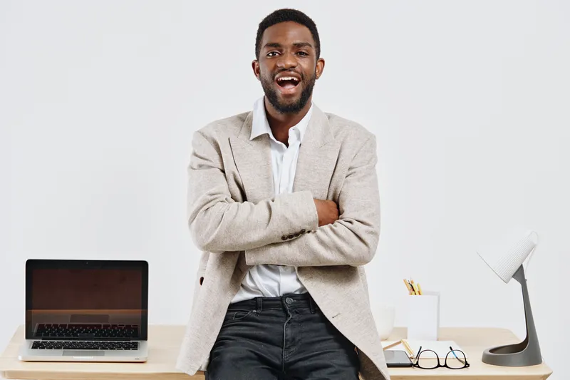 Professional young African American man smiling confidently while standing with arms crossed in modern office, wearing casual beige blazer and white shirt, workspace with laptop and desk accessories, 