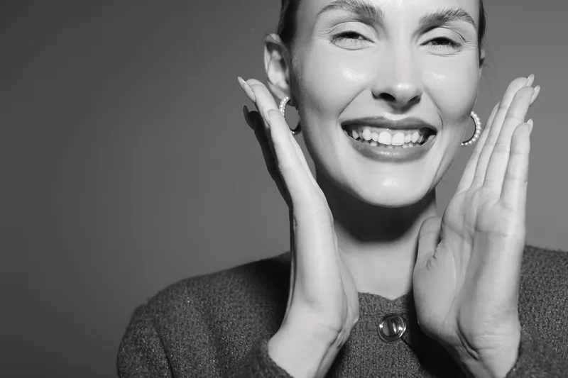 Happy Caucasian woman with fair skin smiles widely and touches face with hands, wearing earrings and textured clothing in studio lighting.