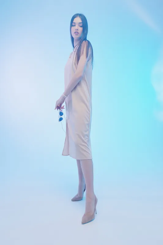 Young woman in a stylish beige dress poses confidently holding sunglasses. Soft blue lighting highlights her beauty and fashion sense in a minimalistic studio setting.