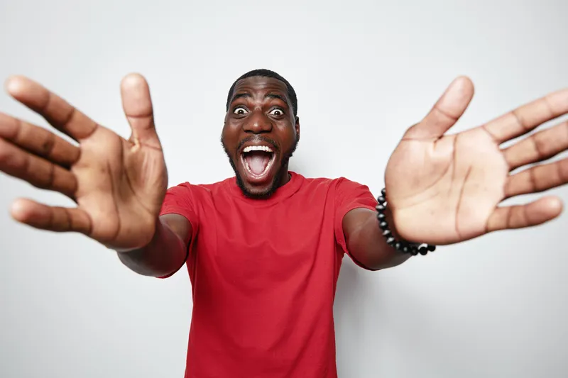 Excited African American man with dark skin wearing red shirt reaching out with open hands and wide smile on white background for happiness and joy concept.