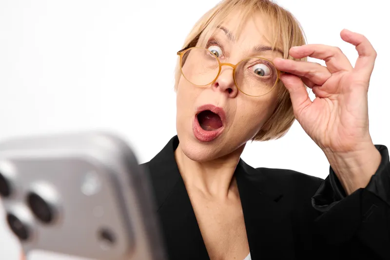 Surprised blonde woman in business suit wearing glasses reacts to smartphone on white background, open mouth and raised fingers, shocked expression, close up dramatic portrait face.