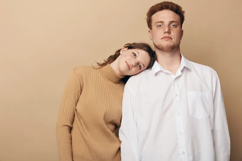 Young couple stands close together against beige background woman in brown sweater leans head on man in white shirt. Concept of relationship support comfort and intimacy.