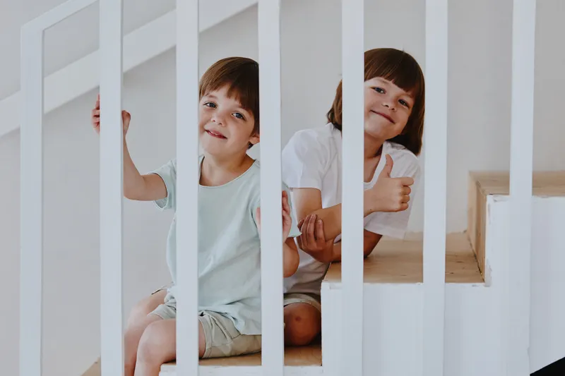Two joyful children posing playfully on the stairs, smiling and giving thumbs up, showcasing happiness in a cozy home setting with bright colors.