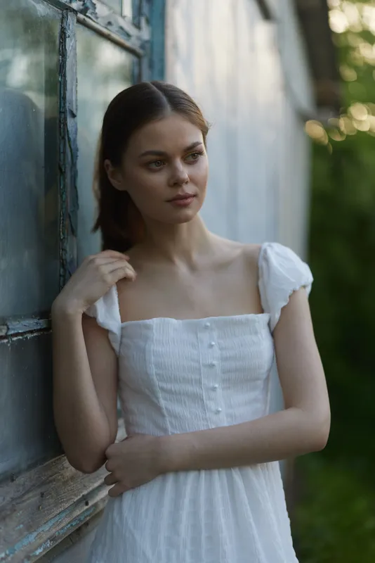 Young woman in a white summer dress standing near an old building, reflecting serenity and nature, embodying the beauty of summer and outdoor portraits