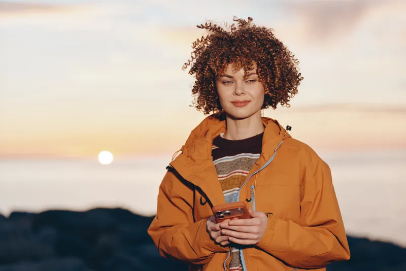 Woman lifestyle portrait outdoors at sunset, curly hair woman holds smartphone in orange jacket, calm smile, warm evening light, distant mountains background, casual travel concept