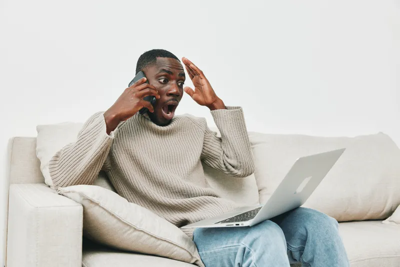 A young Black man in a cozy sweater, shocked while using a laptop and talking on the phone, depicting surprise and excitement in a modern living room setting