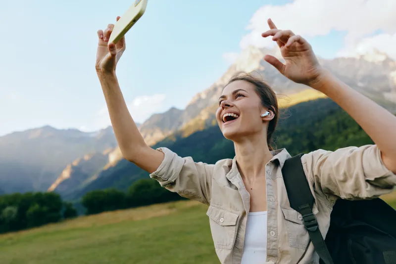 Joyful woman in outdoor setting takes a selfie, smiling with mountains and landscape behind, casual attire, backpack, daytime adventure.