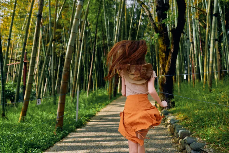 Woman running on a bamboo forest path in summer, orange dress flowing and hair in motion, back view of a person enjoying a sunny nature trail with carefree energy.