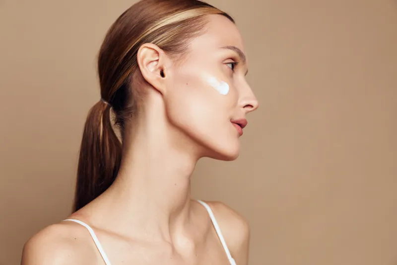 Young adult woman in profile applies white facial cream on her cheek, standing against a neutral beige studio backdrop with soft lighting and a calm, clean beauty look.