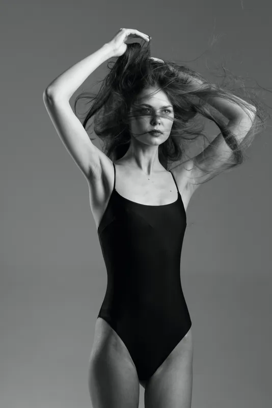 Black and white portrait of a slender woman in a black swimsuit posing with her hands in her hair