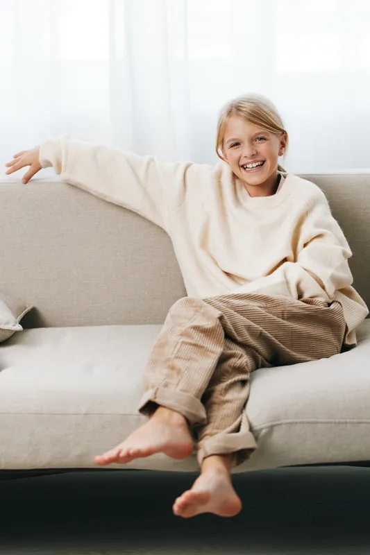 Authentic lifestyle portrait of a cheerful child at home, relaxed on a sofa, wearing a cozy sweater, capturing playfulness, comfort, and everyday school readiness.