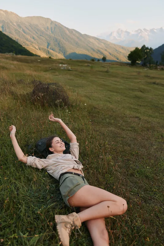 Young woman with long hair lies on green grass in a mountain meadow enjoying a peaceful moment with closed eyes and relaxed arms.