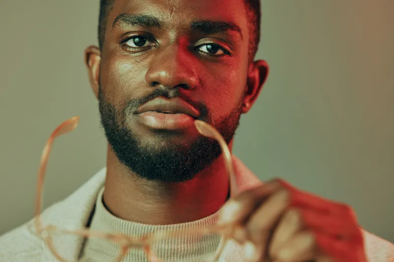 Portrait of a young African man in a stylish sweater holding glasses, conveying confidence and sophistication against a muted background with a warm color palette