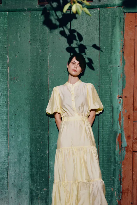 Portrait of a woman in a pale yellow dress standing against a green and orange wall outdoors, bathed in soft sunlight, her calm expression conveys quiet confidence.