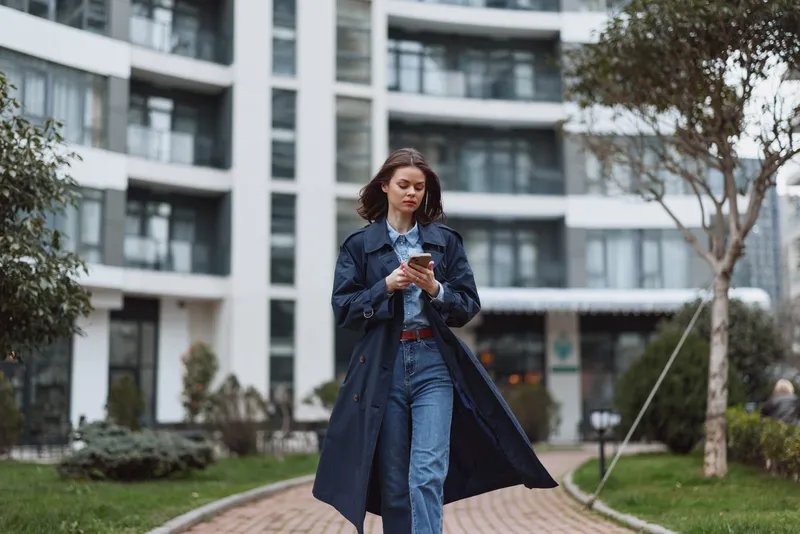 A stylish woman in a long navy coat walks along a campus path, scrolling on her smartphone. Modern buildings frame the scene, illustrating urban lifestyle.