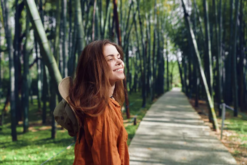Woman smile bamboo forest path hat walking nature portrait happiness outdoor leisure with orange jacket on paved walkway among tall green bamboo trees enjoying peaceful sunlight and fresh air.