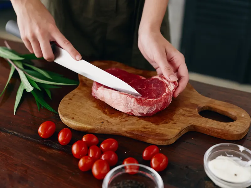 Женщина с ножом в руке нарезает свежее стейковое мясо для жарки на кухне, на столе соль, перец и другие специи, красные помидоры черри и зелень, приготовление ужина