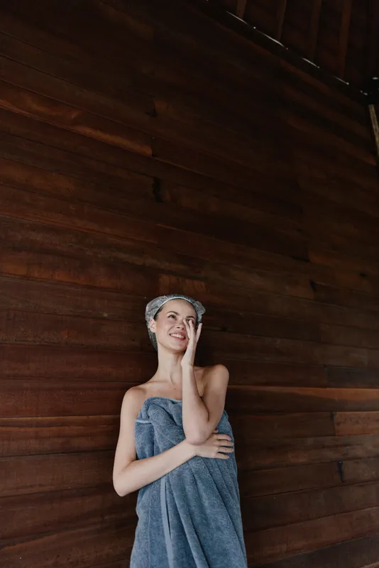 smiling woman wrapped in towel with hair towel standing by dark wooden wall indoors, natural light, relaxed mood, home wellness concept