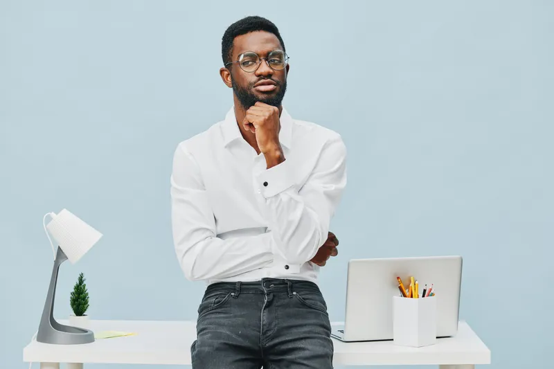 Thoughtful young man with glasses in white shirt standing with crossed arms at modern desk on light blue background, workspace setup, business casual style, professional environment, studio shot. Peop