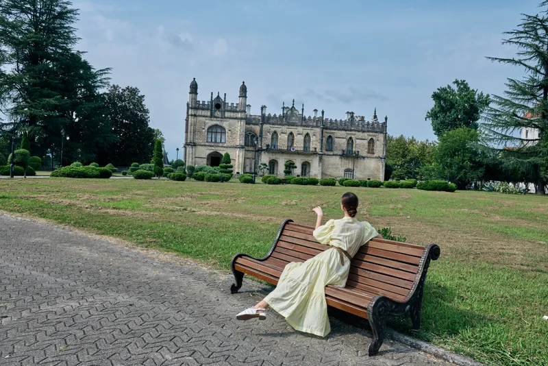 Elegant person in a flowing dress sits on a wooden bench in a sunny park, with a historic castle in the background, creating a serene and timeless outdoor scene for calm moments today.