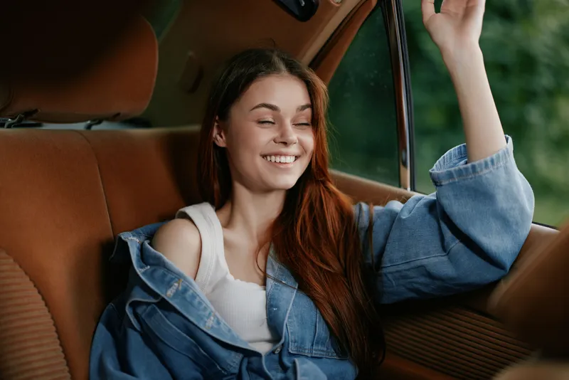 Happy young woman with long hair in denim jacket enjoying the drive in a vintage car, smiling at the sunlight shining through the window.