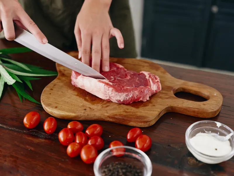 Женщина с ножом в руке нарезает свежий стейк из мяса для жарки на кухне, на столе соль, перец и другие специи, красные помидоры черри и зелень, приготовление ужина