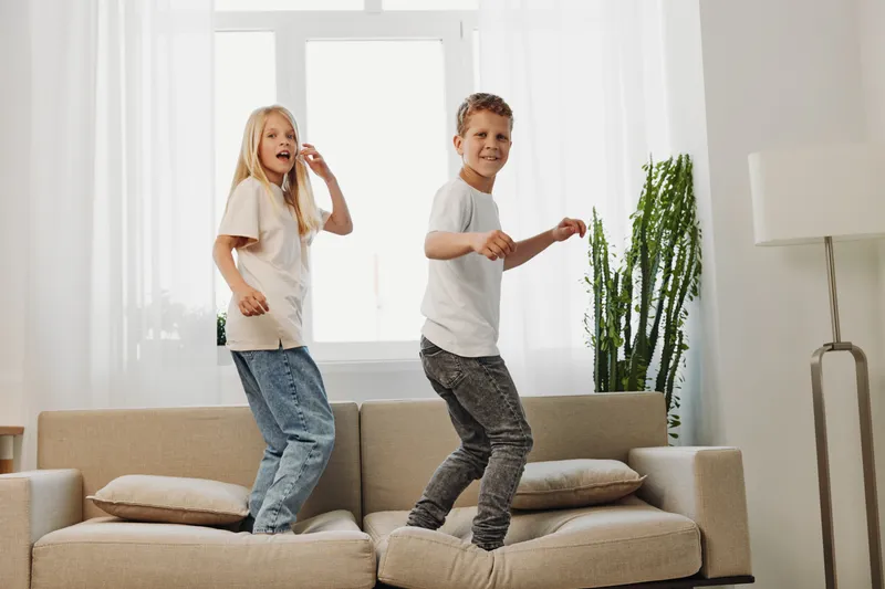 Two happy children jump on a beige sofa in a bright living room with natural light. The boy and girl wear casual clothes and enjoy playful moments at home with plants near the window.
