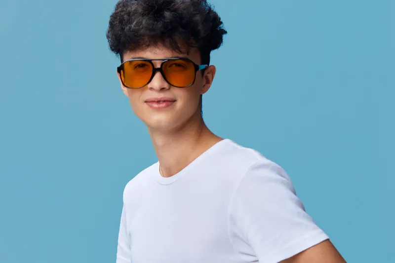 Young man wearing orange sunglasses and white t-shirt posing confidently against a plain blue background with casual hairstyle and relaxed expression.