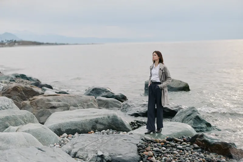 Young woman stands on rocky shore looking at sea horizon. Calm water and cloudy sky create peaceful mood. Casual outfit with jacket and trousers suits outdoor nature concept.