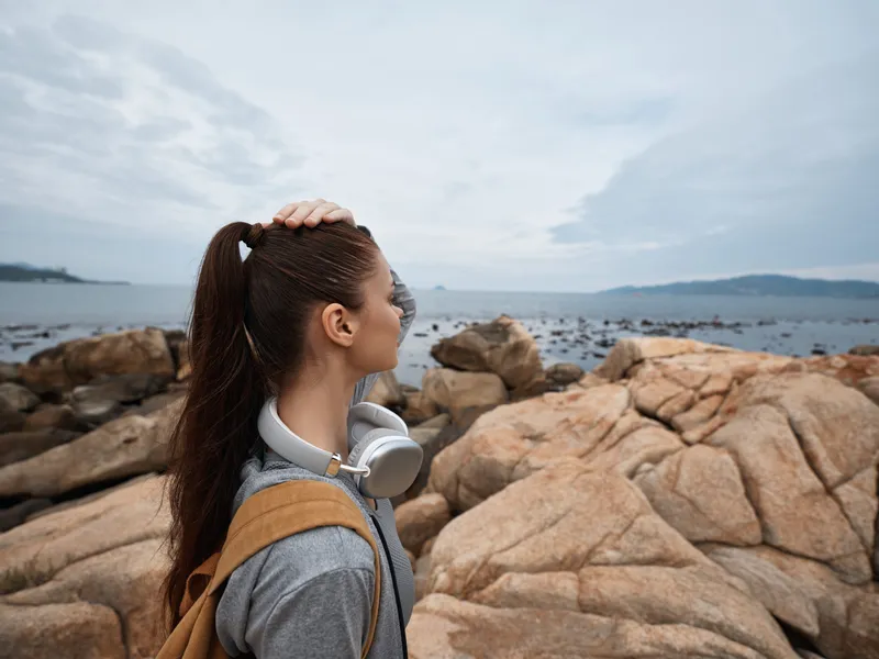 Young woman exploring the beach, wearing headphones and a backpack, looking at the ocean, with a calm expression and a cloudy sky backdrop, captures wanderlust
