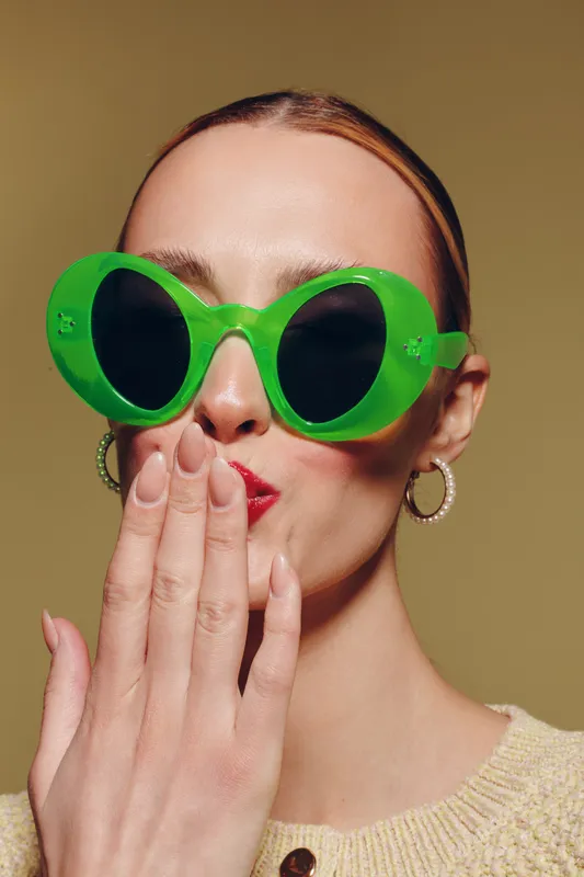 Woman with green sunglasses and red lipstick covers mouth with hand. Close-up portrait on beige background. Concept of fashion, style, and playful expression.