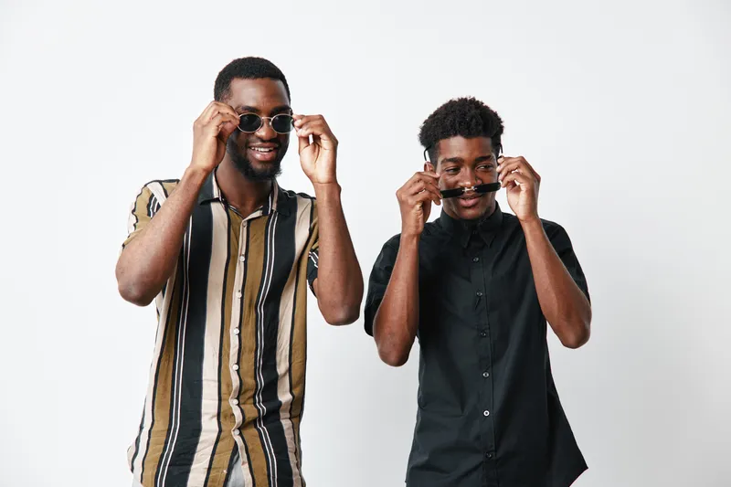 Two young black men with dark skin wearing sunglasses and casual shirts smiling and posing together in studio with white background for friendship and style concept.