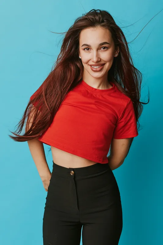 Young woman with long hair wearing red cropped t shirt and black pants smiling and posing against blue background in studio. Casual style and confident expression.