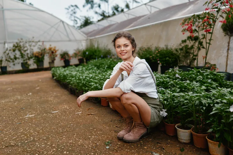 Young woman in a greenhouse, wearing casual attire, showcasing a joyful expression, surrounded by vibrant plants highlighting a connection to nature and freshness