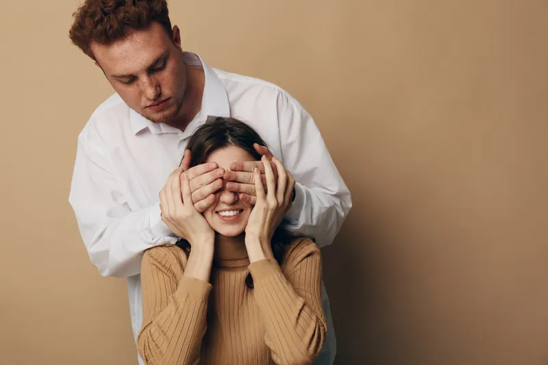 Young man covers eyes of smiling woman from behind in cozy beige sweater. Intimate moment of surprise and affection captured in warm studio with neutral background. Concept of love.
