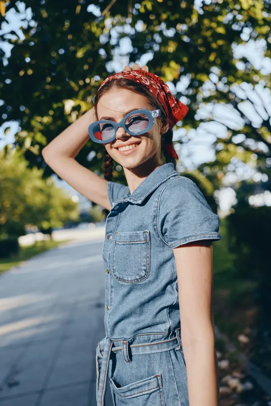 Lifestyle woman in modern boho-western denim street style wearing heart-shaped sunglasses and red bandana, smiling joyfully outdoors in natural sunny setting.