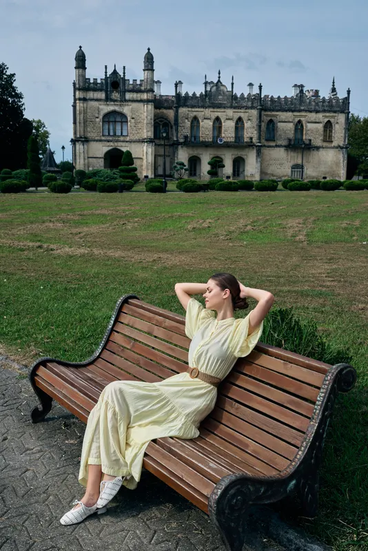 Portrait of a woman in a pale yellow dress relaxing on a wooden bench in a formal garden with a historic castle in the background creating a serene and elegant atmosphere.