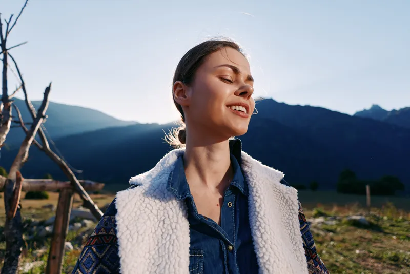 Woman portrait in mountains nature at golden sunlight, smiling and happy outdoors with casual jacket and wool vest, scenic landscape background and peaceful rural vibe during sunset.