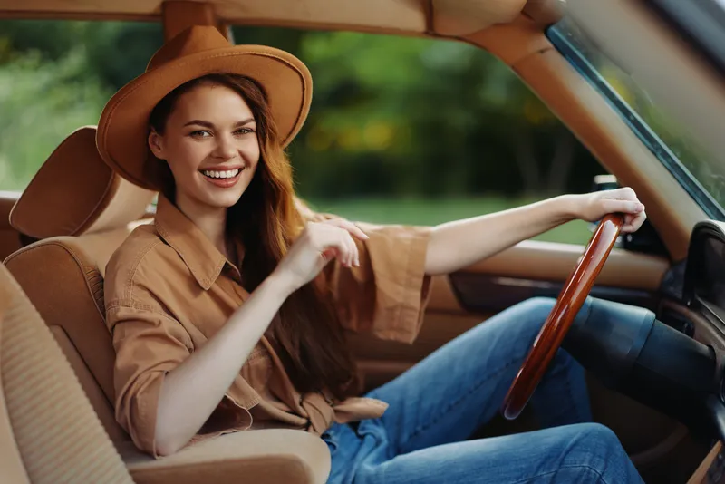 Happy young woman in a car wearing a stylish hat and casual clothing, showcasing a joyful expression in a natural green background.