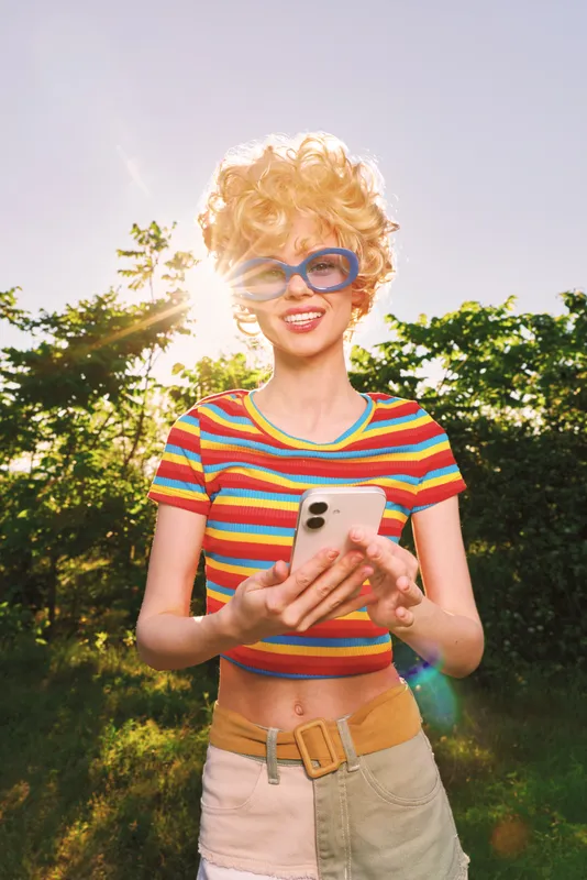 Cheerful woman outdoors, wearing a striped top, using a smartphone, sunlit moment with soft breeze, casual pose, bright colors, lush greenery background, lifestyle portrait