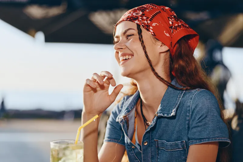 smiling woman with braided hair wearing red bandana and denim jacket enjoying drink outdoors in sunny weather with relaxed joyful expression