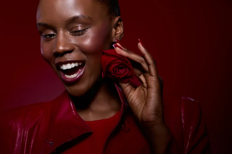 Closeup of happy dark skinned African woman winking and holding red rose near face wearing red jacket and shirt. Concept for beauty, joy, romance, and emotional expression with vibrant colors.