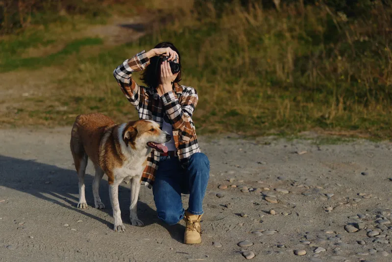 Dog, woman, outdoors, companionship. A woman kneels beside a dog on a gravel beach, plaid shirt, calm moment, sunlit scenery, candid outdoor portrait capturing companionship and nature in a peaceful