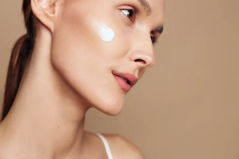 Young woman applies white facial cream on her cheek in a close up studio shot with a soft beige background, conveying skincare and self care.