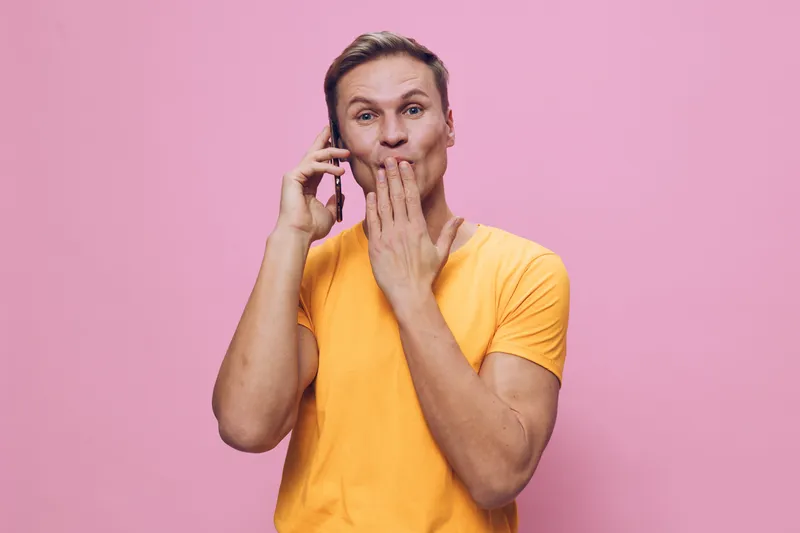 Playful young man with surprised expression talking on mobile phone isolated on pink background. Casual yellow t-shirt, fun mood, communication, lifestyle concept, studio portrait.