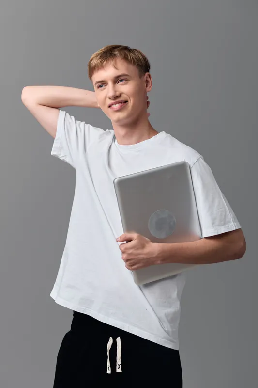 Man in a casual white tee holding a laptop, smiling in a studio setting, conveying youth energy, learning and technology readiness for education, remote work, or creative projects